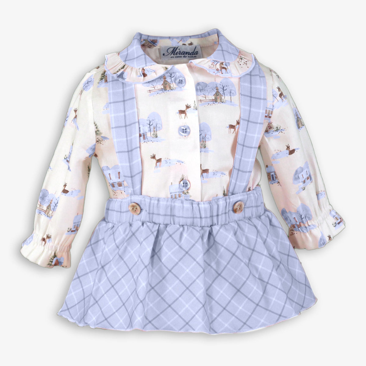 A baby girl's long-sleeve dress with a winter print and a light blue plaid skirt. The top features a white background with a light blue plaid collar and cuffs, and a central panel with a winter scene print including trees, houses, and deer.